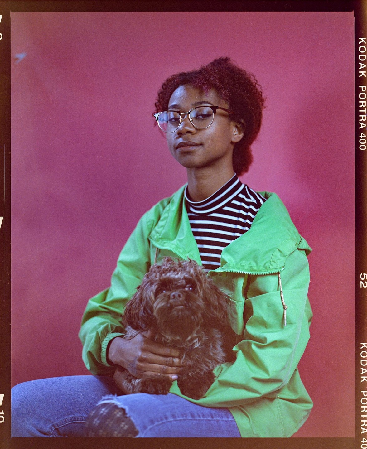 A woman wearing glasses and a green jacket holds a small dog against a pink background.