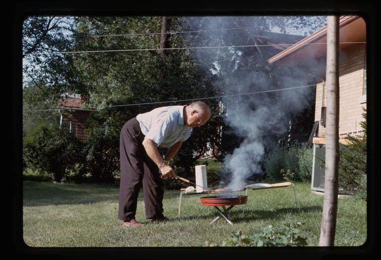 A man wearing glasses bends over a smoking barbecue grill in a grassy yard.