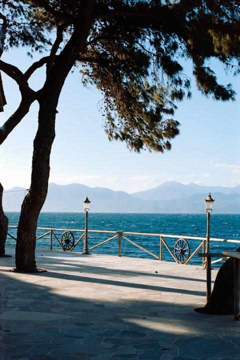 A tree casts a shadow on a stone pier next to a body of water with mountains in the distance.