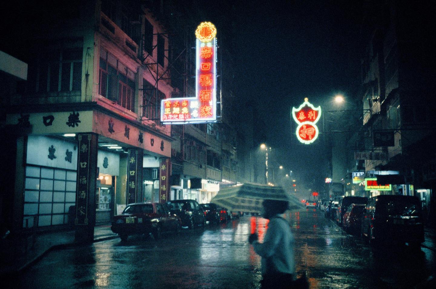 A person with an umbrella walks down a wet street lined with cars and buildings with neon signs at night.