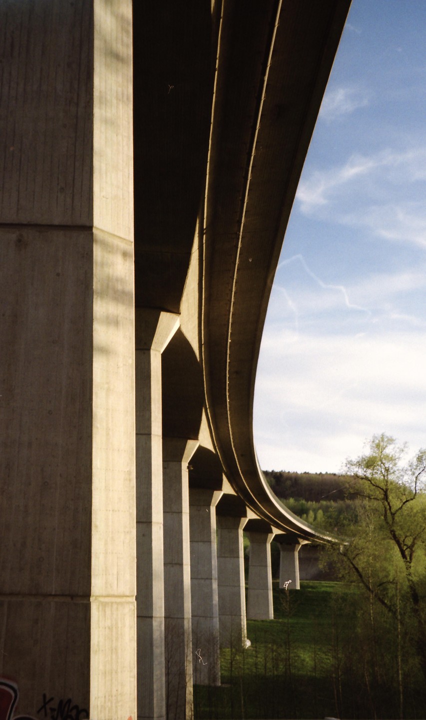 A concrete highway overpass curves over a green hillside with trees under a blue sky.