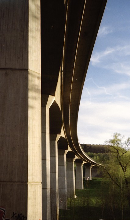A concrete highway overpass curves over a green hillside with trees under a blue sky.