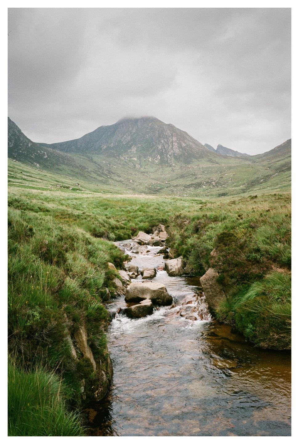 A stream flows over rocks through a grassy valley with mountains under a cloudy sky.
