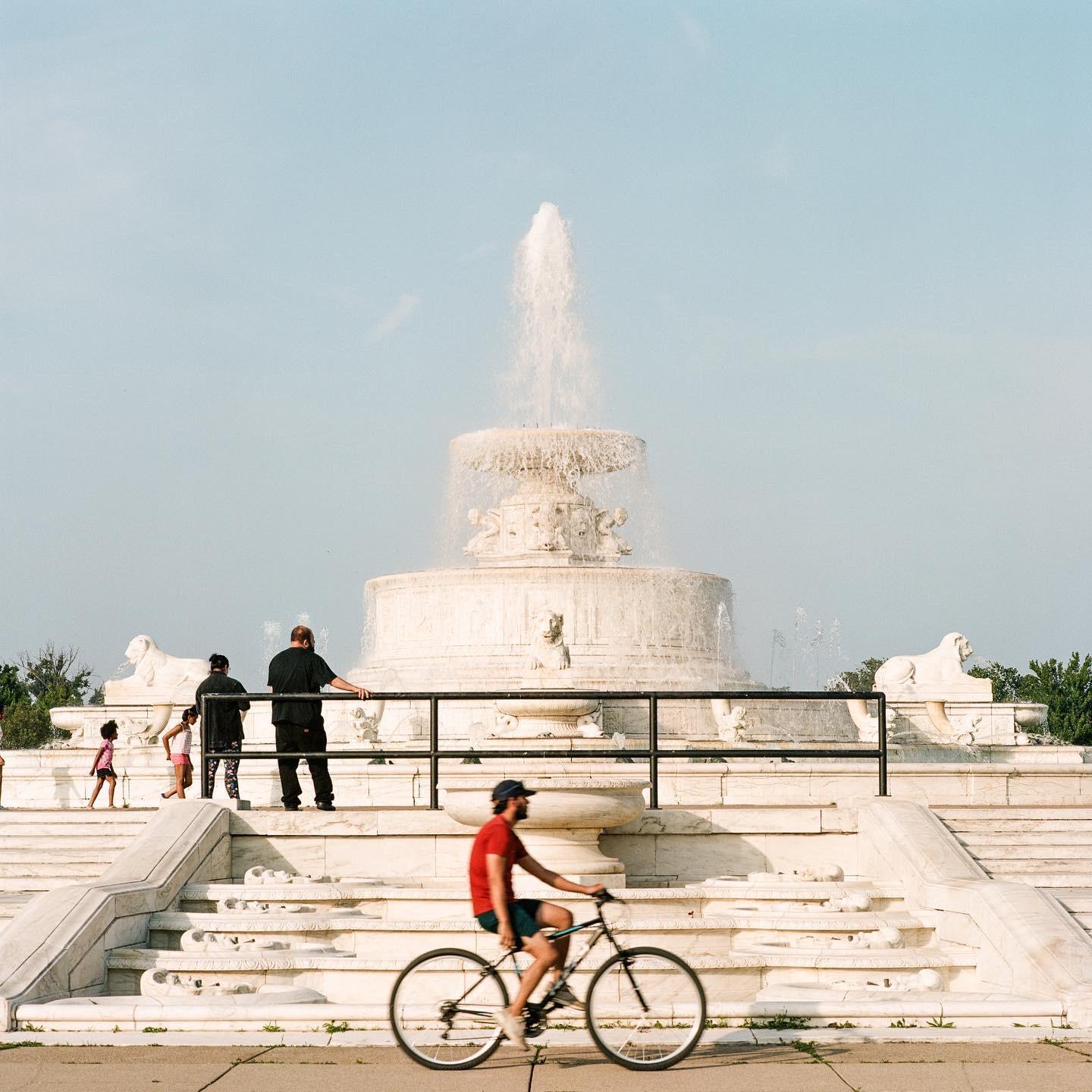 A man on a bicycle rides past a large white fountain with people standing on its steps.