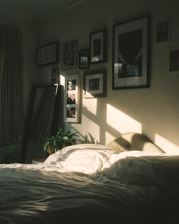 Sunlight streams into a bedroom, illuminating a bed with white linens and framed pictures on the wall.