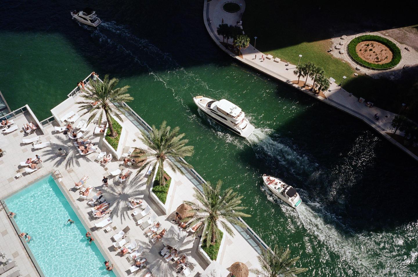 An aerial view shows boats on a canal next to a building with a swimming pool and people sunbathing.