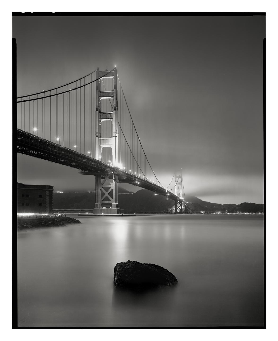The Golden Gate Bridge is illuminated at night with a large rock in the foreground and a building on the left.