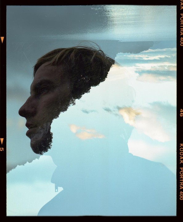 A man's profile is double exposed with a landscape of water, trees, and clouds.