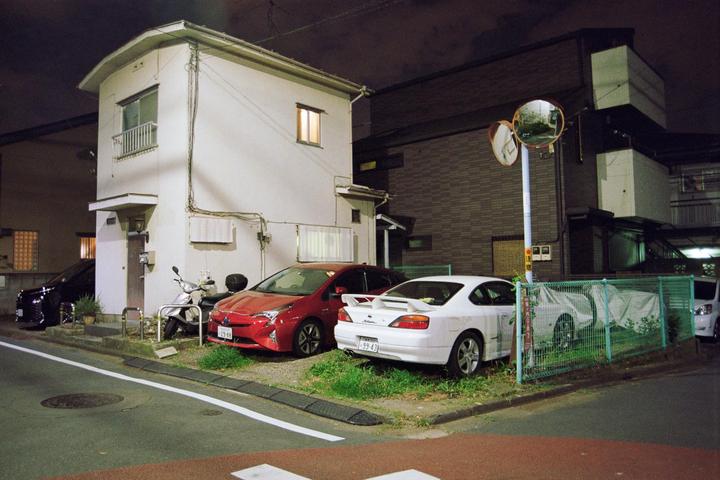A white house with illuminated windows stands next to a street with parked cars and a scooter at night.