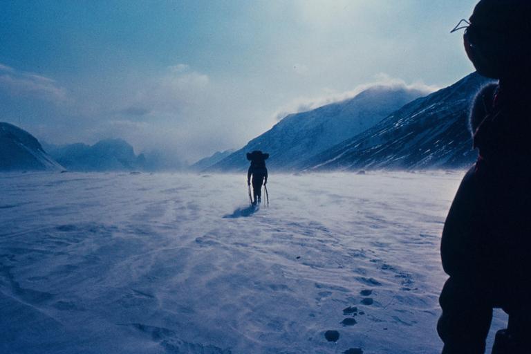 A person with a backpack and trekking poles walks across a snowy, windswept plain with mountains in the background.