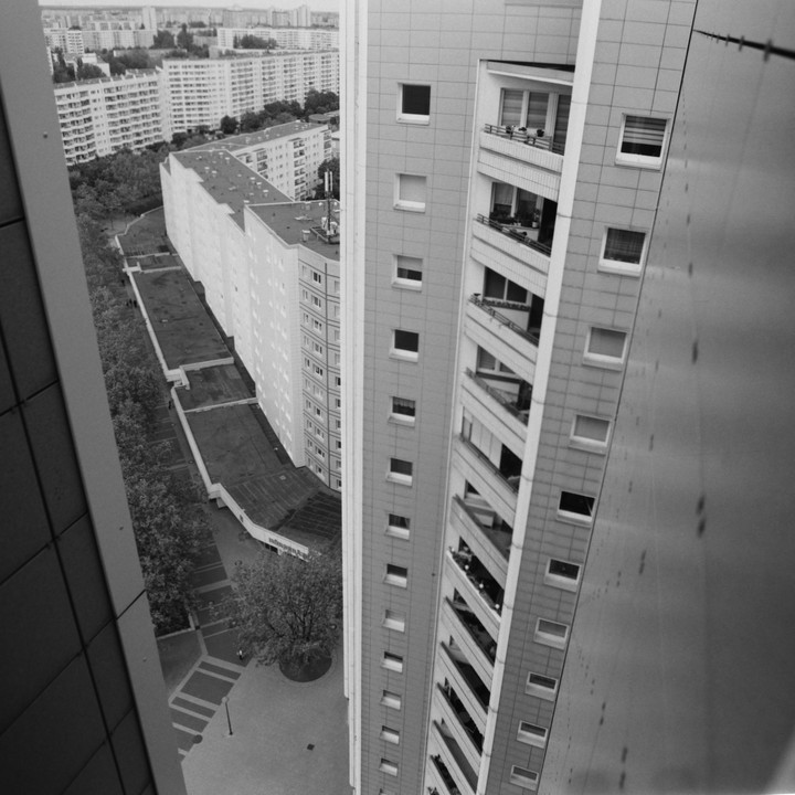 A high-angle view shows a tall building with balconies next to a street with trees and other buildings in the background.
