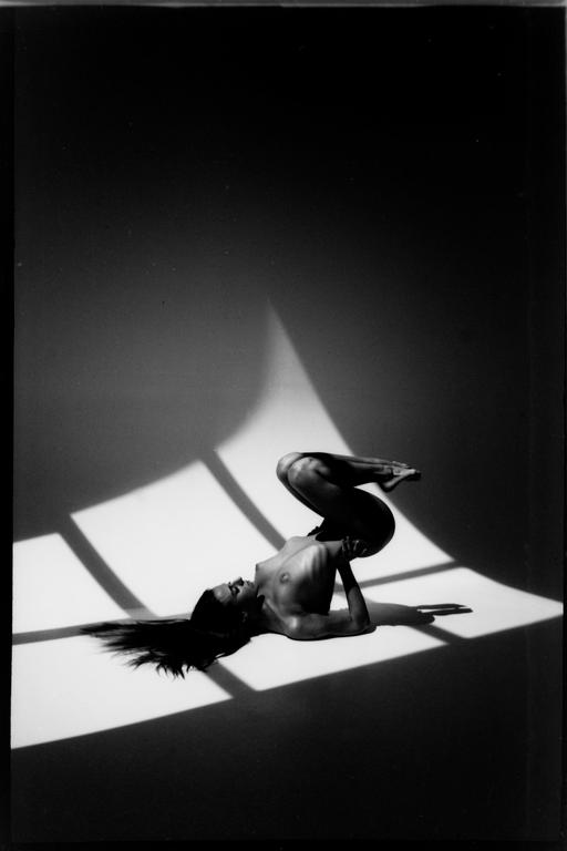 A woman lies on her back with her legs bent and raised, illuminated by window light and shadow.