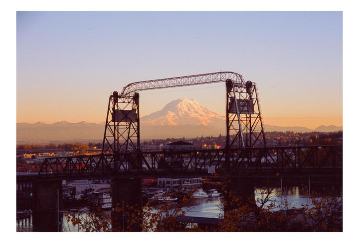 A snow-capped mountain is visible through the opening of a bridge over a body of water with boats.