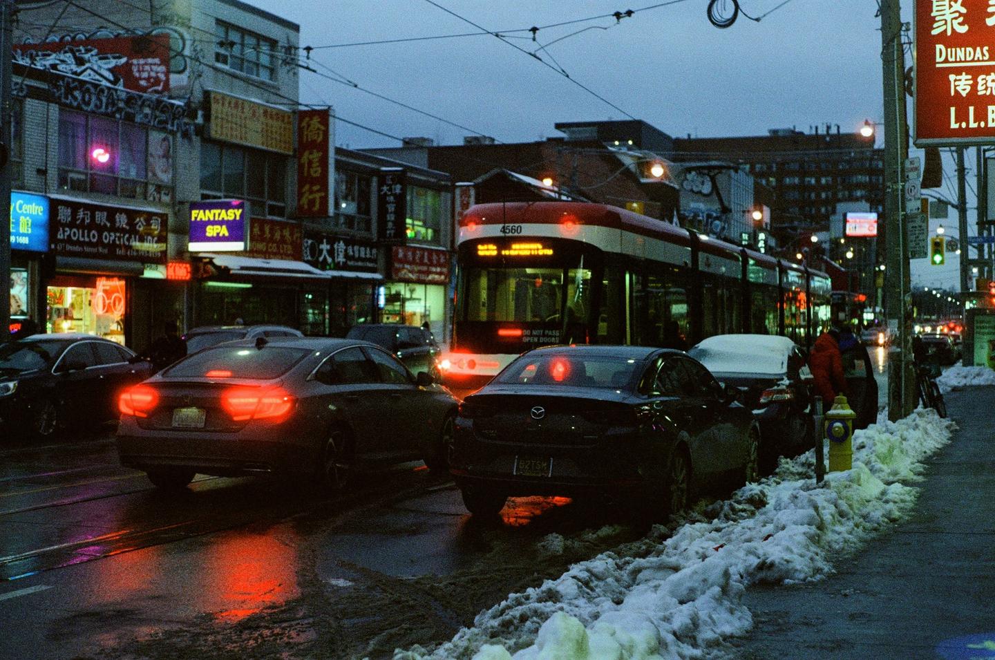 A streetcar drives down a city street at night, with cars parked on the side of the road and snow on the ground.