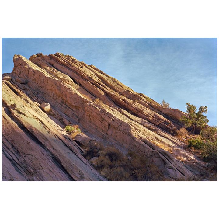 A large rock formation with diagonal strata is illuminated by sunlight under a blue sky.