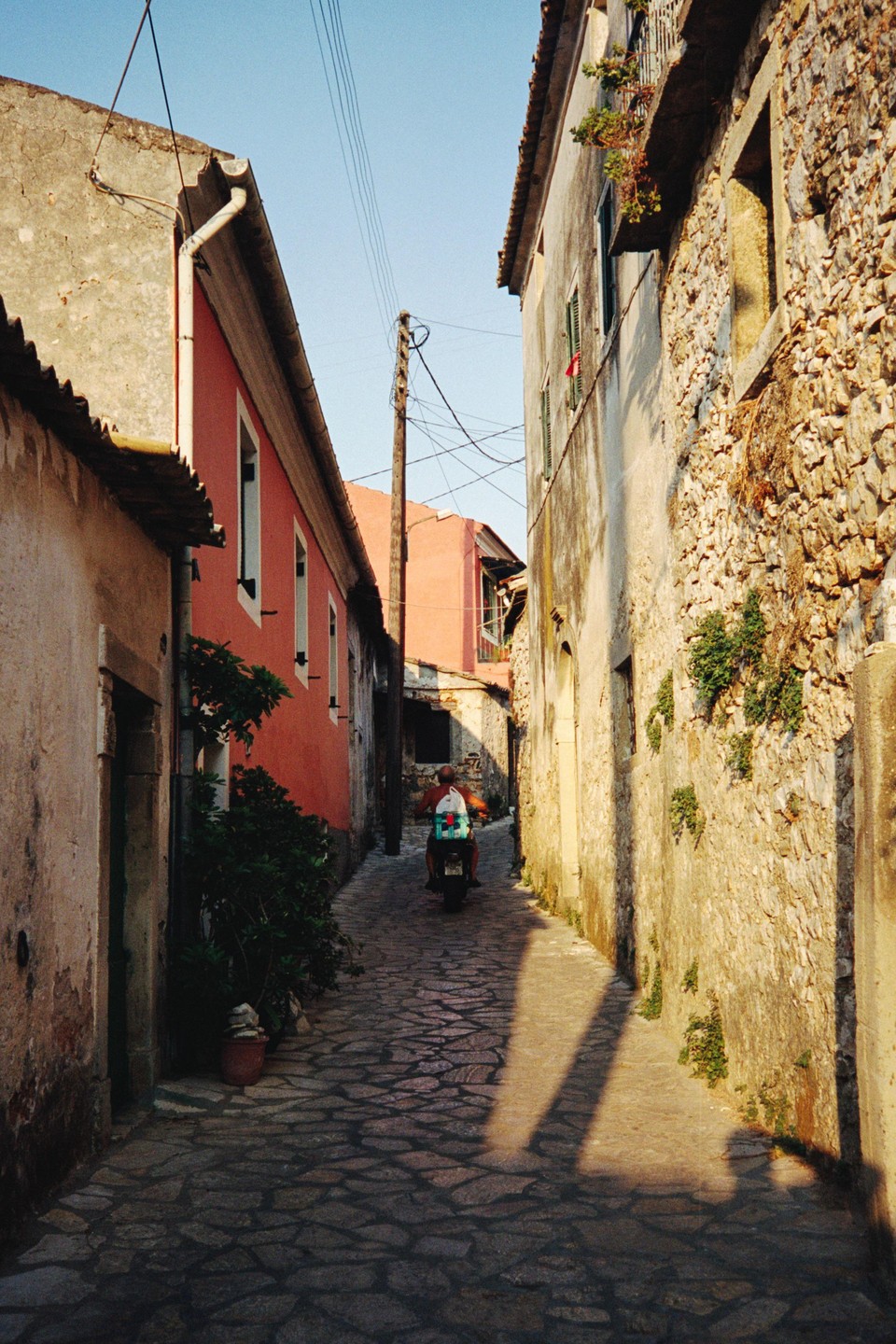 A man rides a scooter down a narrow, cobbled street between old buildings in Greece.