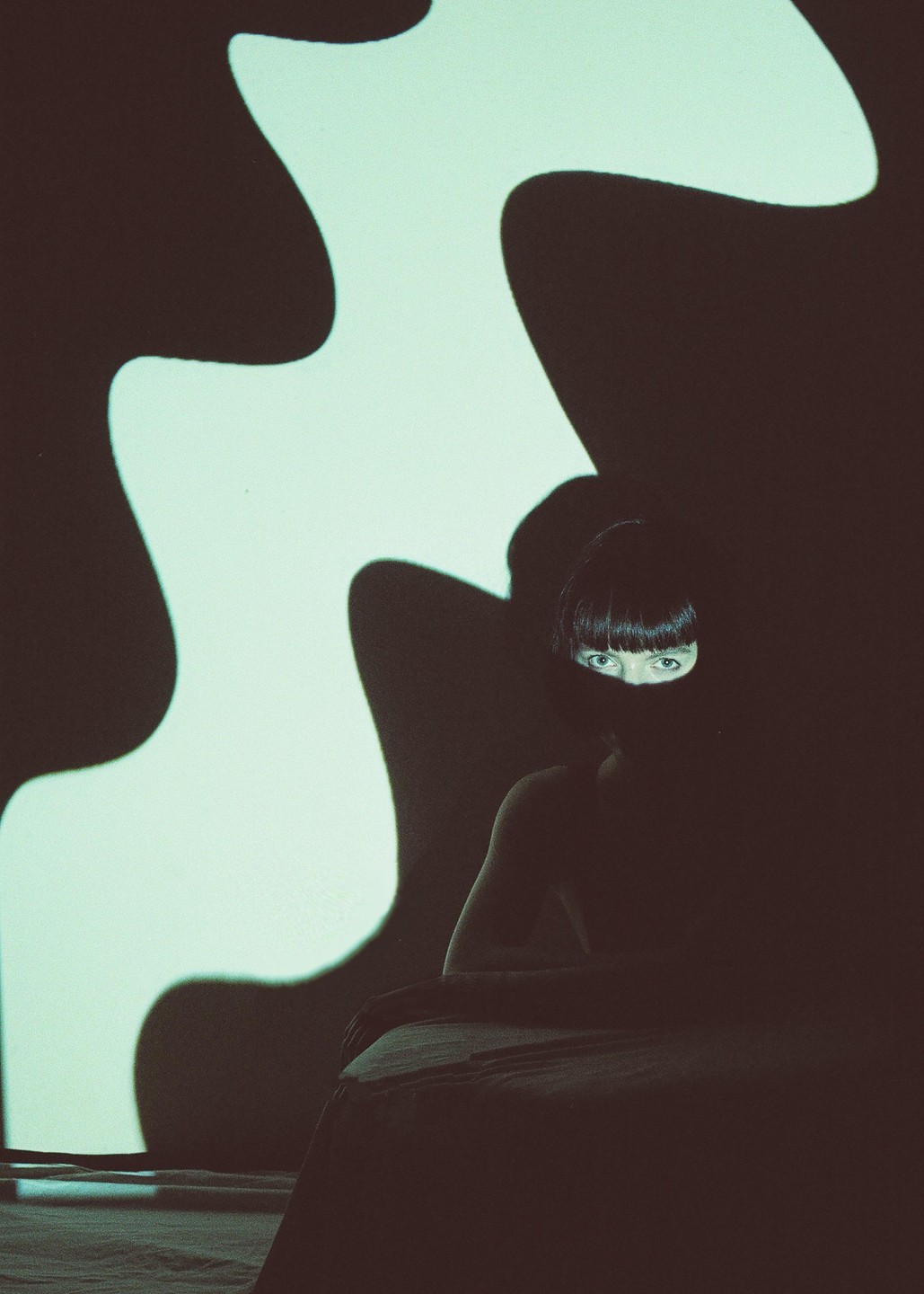 A woman with bangs sits in shadow with only her eyes visible, next to a wall with a wavy light projection.