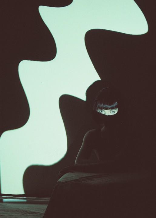 A woman with bangs sits in shadow with only her eyes visible, next to a wall with a wavy light projection.