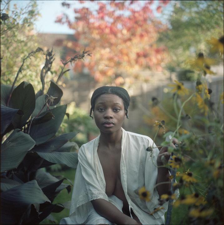 A woman in a white robe sits in a garden with large green leaves and yellow flowers.
