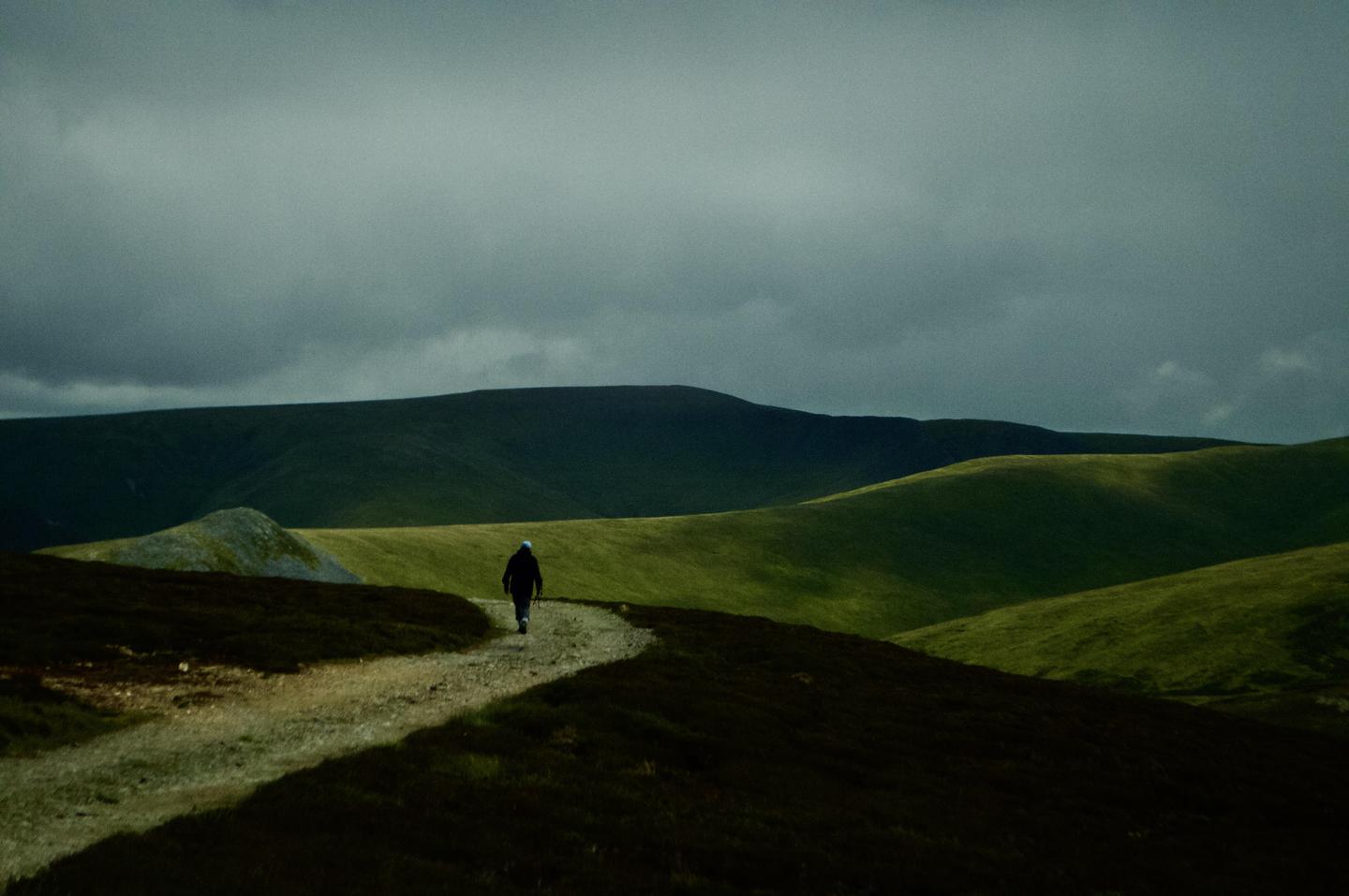 A person walks along a winding path through rolling hills under a cloudy sky.