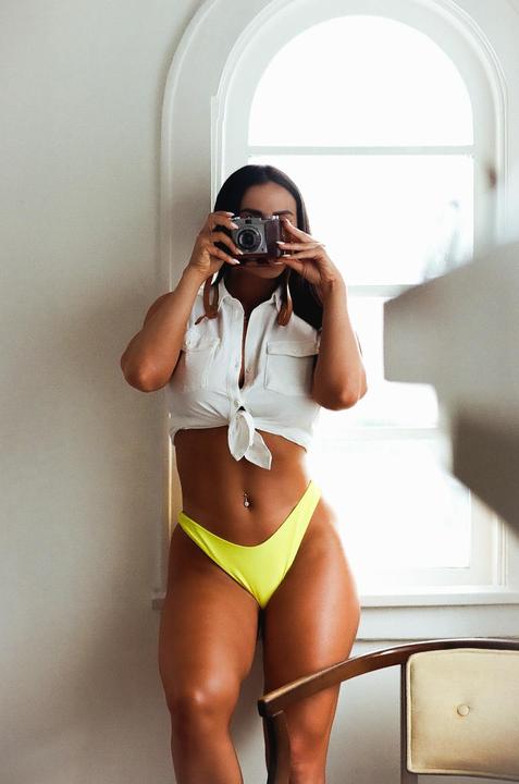 A woman in a white shirt and yellow bikini bottom holds a camera to her face, standing in front of an arched window.