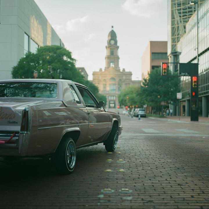 A pink lowrider car is parked on a brick street with a clock tower building in the background.