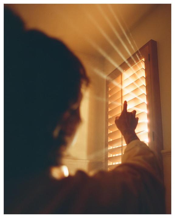 A person reaches out to touch light rays streaming through window blinds.