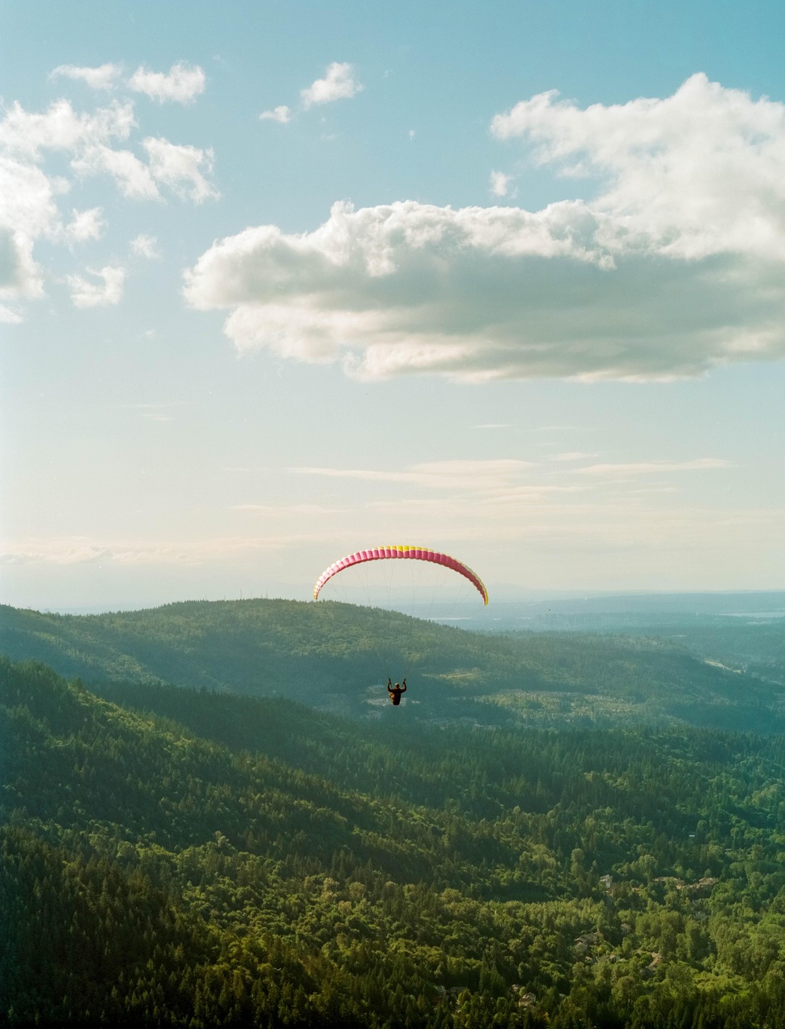 A paraglider soars over a vast expanse of green forest under a partly cloudy sky.