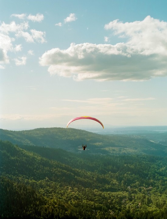 A paraglider soars over a vast expanse of green forest under a partly cloudy sky.