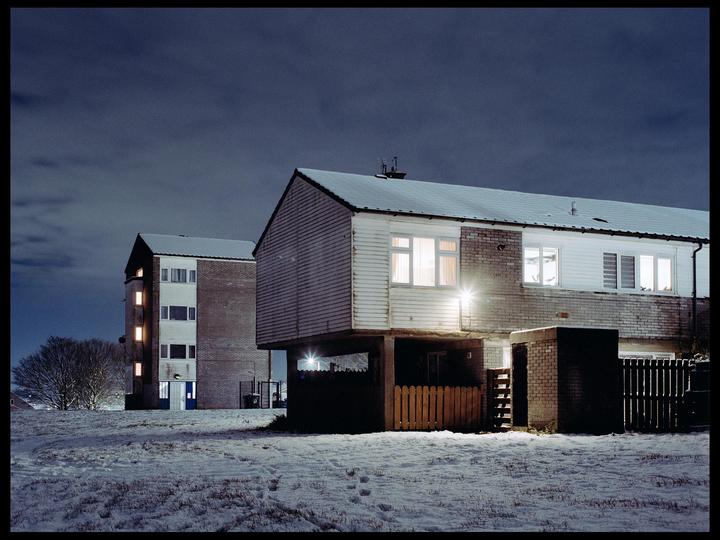 Two buildings with snow-covered roofs stand in a snowy field under a dark sky at night.