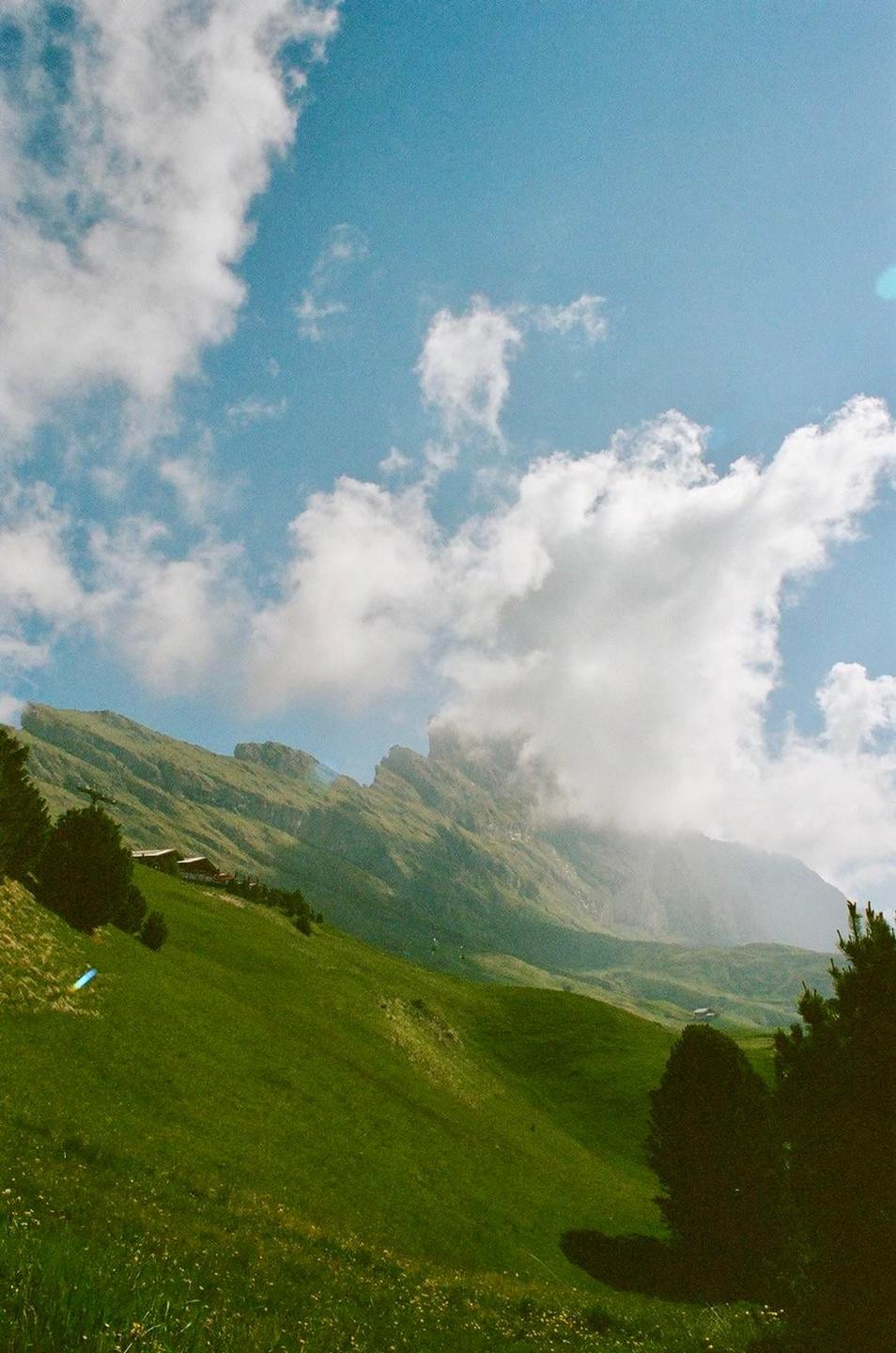 A green mountain slope with scattered trees and buildings is visible under a blue sky with white clouds.