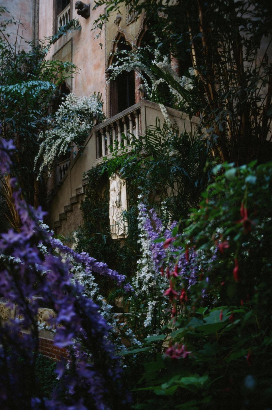 A stone building with a balcony and stairs is surrounded by lush foliage and colorful flowers.