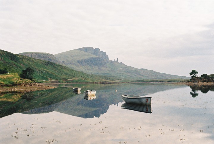 Three small boats float on a calm lake with mountains and their reflections in the background.