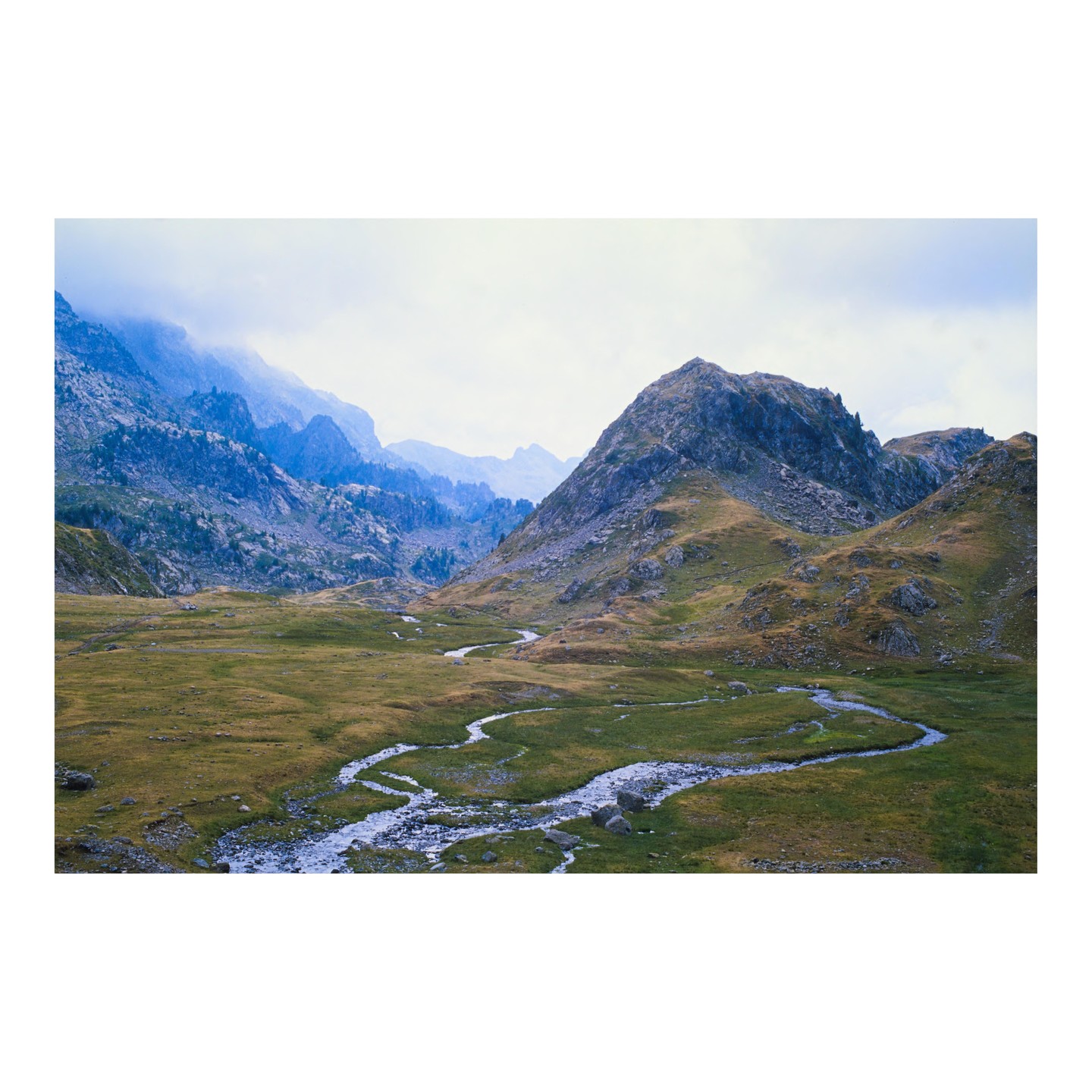 A winding river flows through a grassy valley surrounded by mountains under a hazy sky.