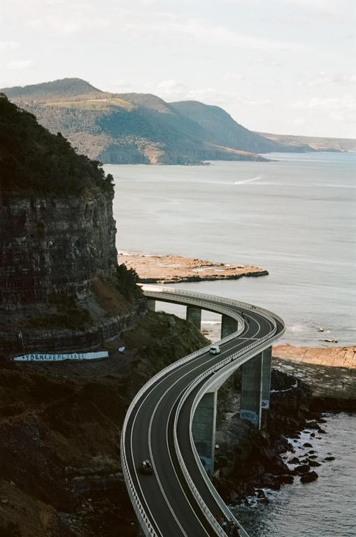 A winding coastal road bridge extends over the ocean, with mountains in the background.