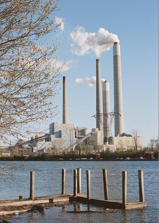 A power plant with multiple smokestacks emitting smoke is visible across a body of water, with a small wooden dock in the foreground and tree branches on the left.