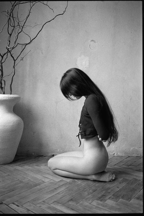 A woman with long hair kneels on a wooden floor next to a large vase with bare branches.