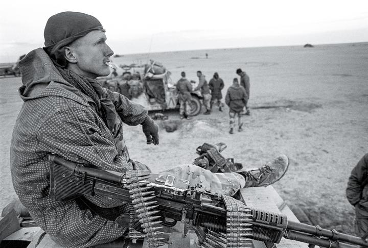 A man in military fatigues with a cigarette in his mouth sits next to a machine gun with a belt of ammunition, looking out over a desert landscape where other soldiers are gathered.