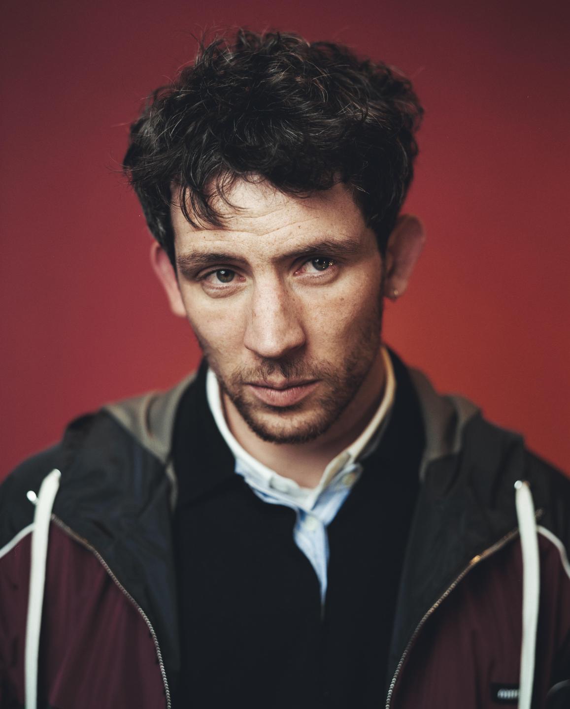 A man with dark, curly hair and a beard looks directly at the viewer against a red background.