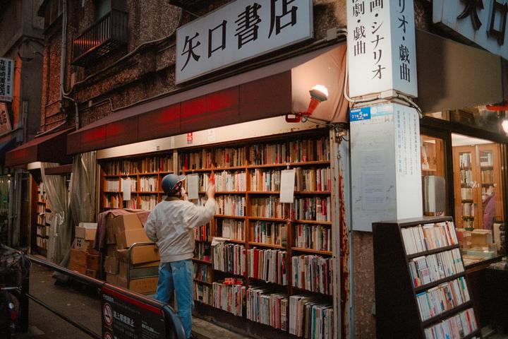 A man wearing a helmet stands in front of a bookstore with shelves of books lining the exterior wall.