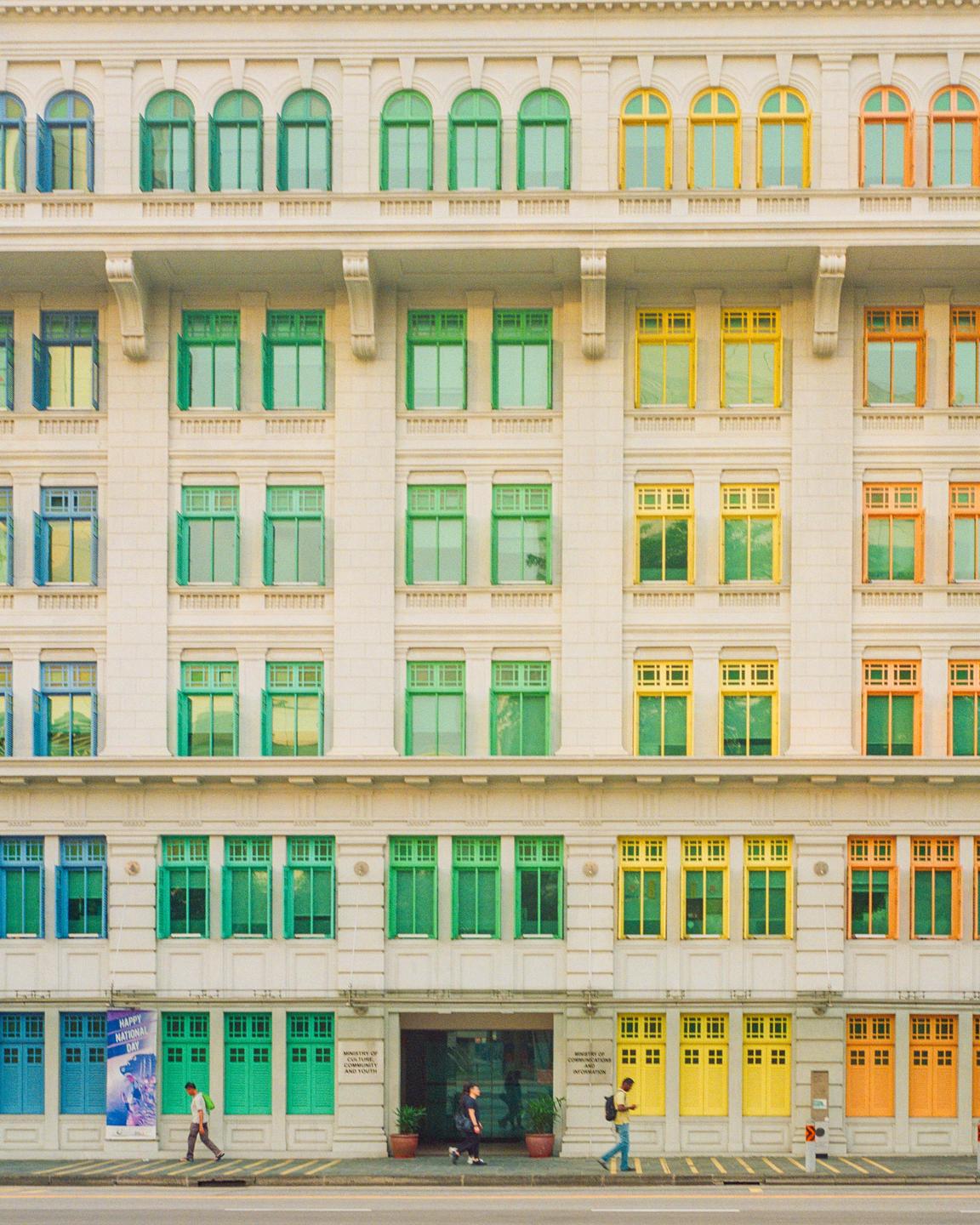 A large building with many colorful windows and doors stands next to a street with people walking on the sidewalk.