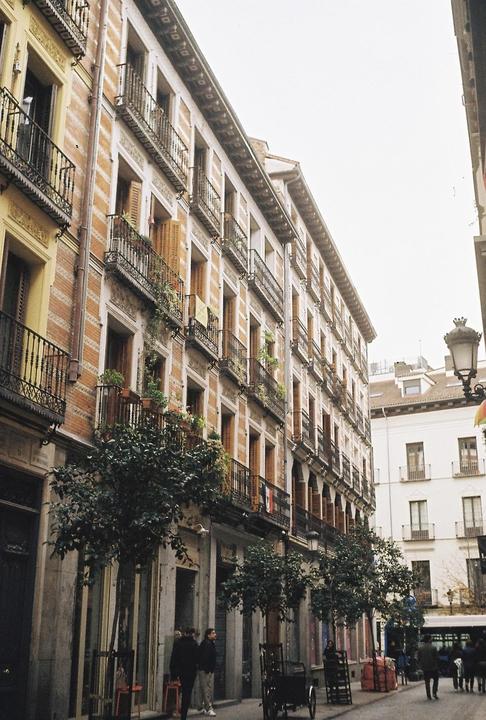 A street in Madrid is lined with tall buildings featuring balconies and trees, with people walking along the sidewalk.