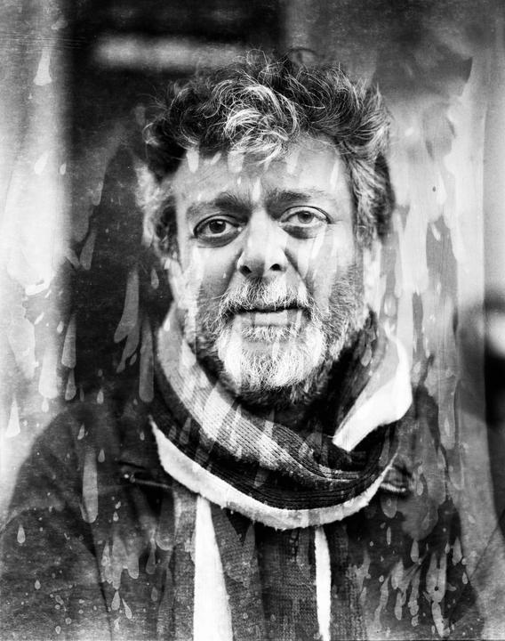 A man with a beard and a striped scarf looks directly at the viewer, with water damage visible on the image.