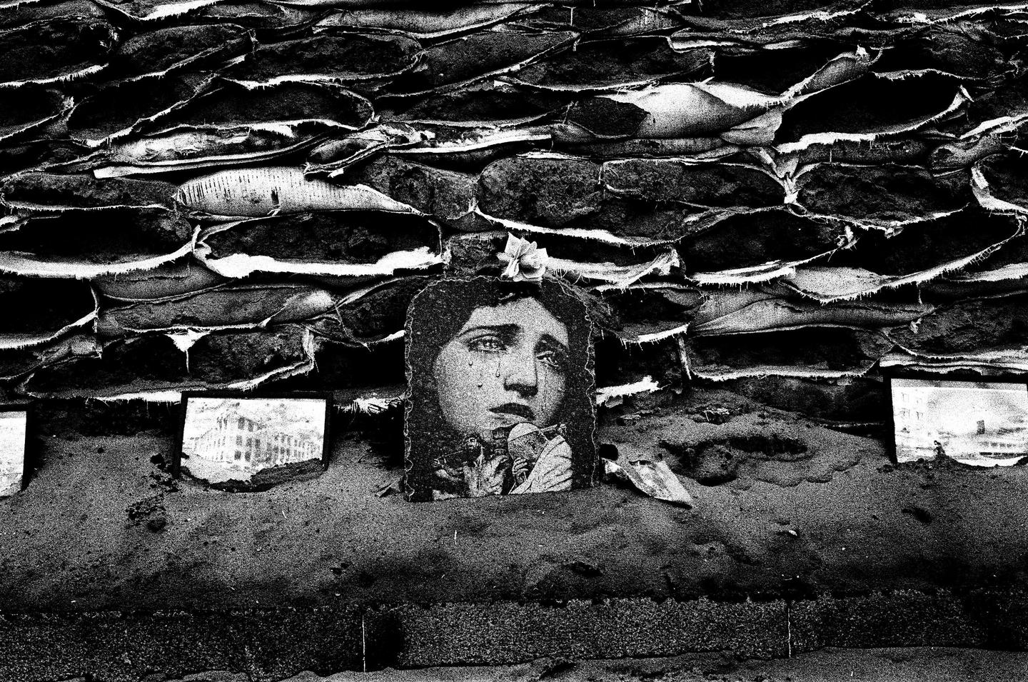 A cutout of a crying woman's face is placed against a wall of sandbags, with a small flower on top of her head.
