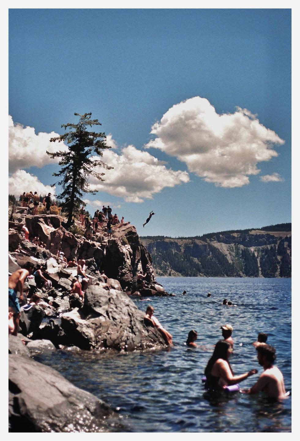 A person jumps from a rocky cliff into a lake where other people are swimming, with mountains in the background under a blue sky with clouds.