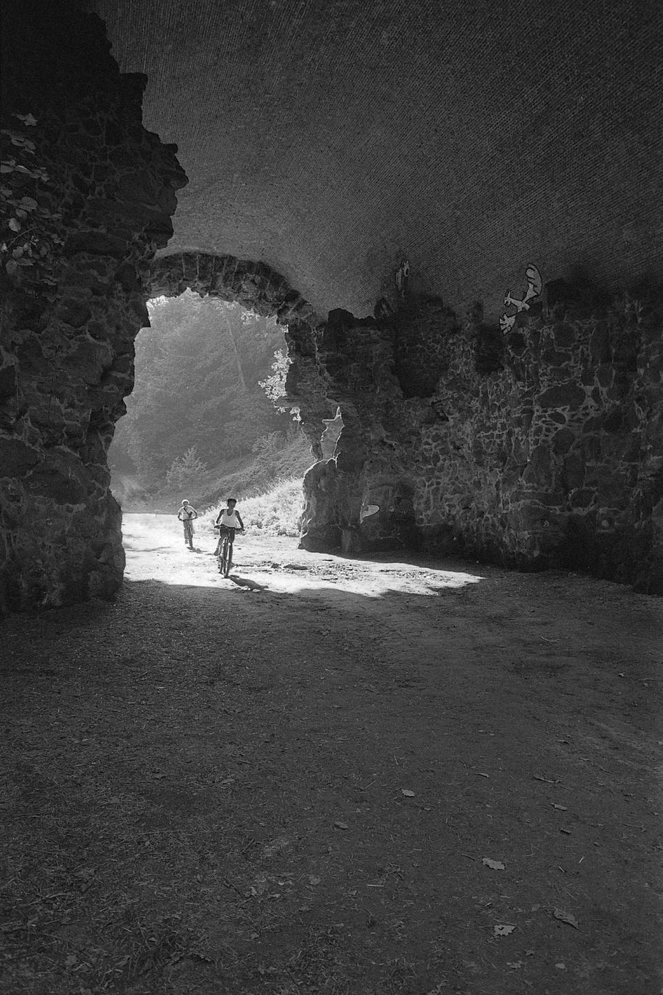 Two children are visible in a sunlit tunnel, one riding a bicycle and the other walking behind.