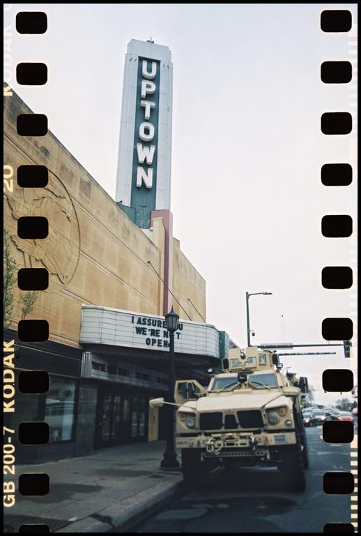 A military vehicle is parked on a street in front of a movie theater with a tall "Uptown" sign.
