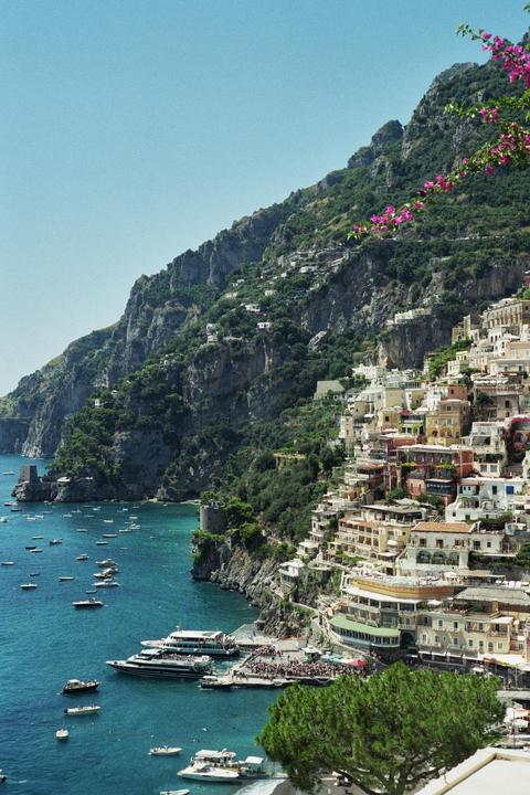 A coastal town with colorful buildings is nestled into a steep, green mountainside overlooking a bay filled with boats.