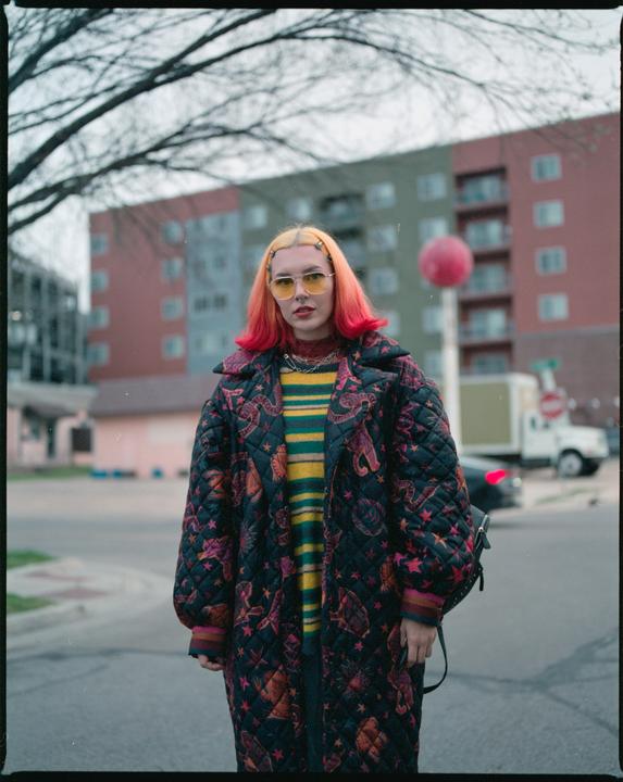 A woman with bright orange hair and yellow sunglasses stands on a street in front of a building.