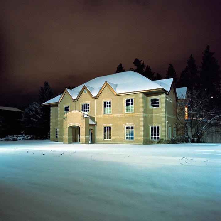 A large brick house with a snow-covered roof stands in a snowy field at night, with trees in the background.